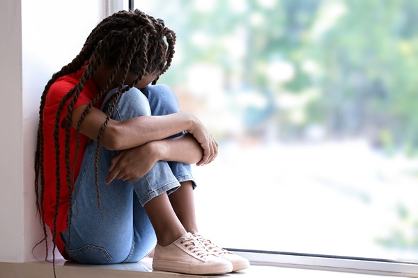 Young girl sitting by a window with her head down and arms wrapped around her knees, appearing sad or distressed. (366077404)
