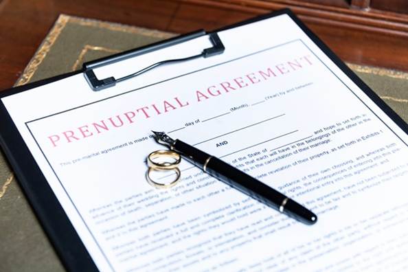 A Prenuptial Agreement document on a black clipboard, with a fountain pen and two gold wedding rings resting on the paper. (740756933)