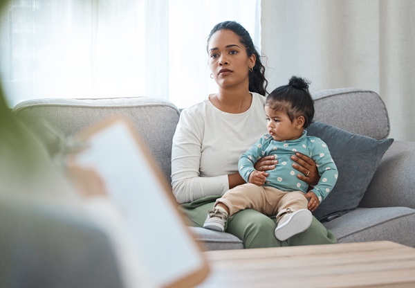A concerned mother sitting on a grey sofa holding her young child while looking toward a DCPP interviewer holding a clipboard during a formal investigation in New Jersey.