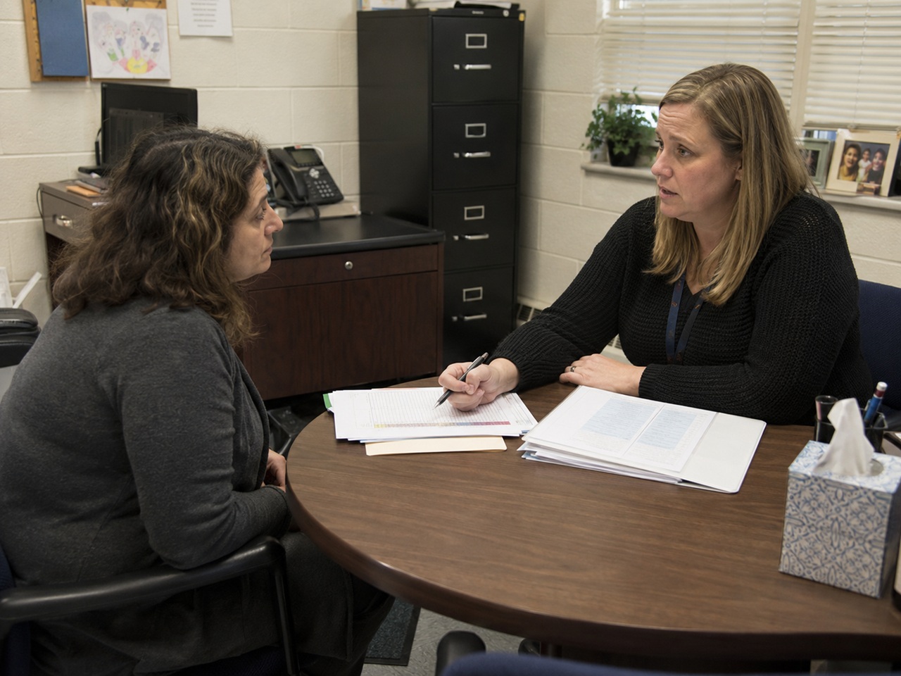 A visibly anxious woman sits across a round table from a caseworker holding a pen and case documents in a sparse government office, depicting a New Jersey DCPP educational neglect investigation interview between a parent and a Division of Child Protection and Permanency worker.